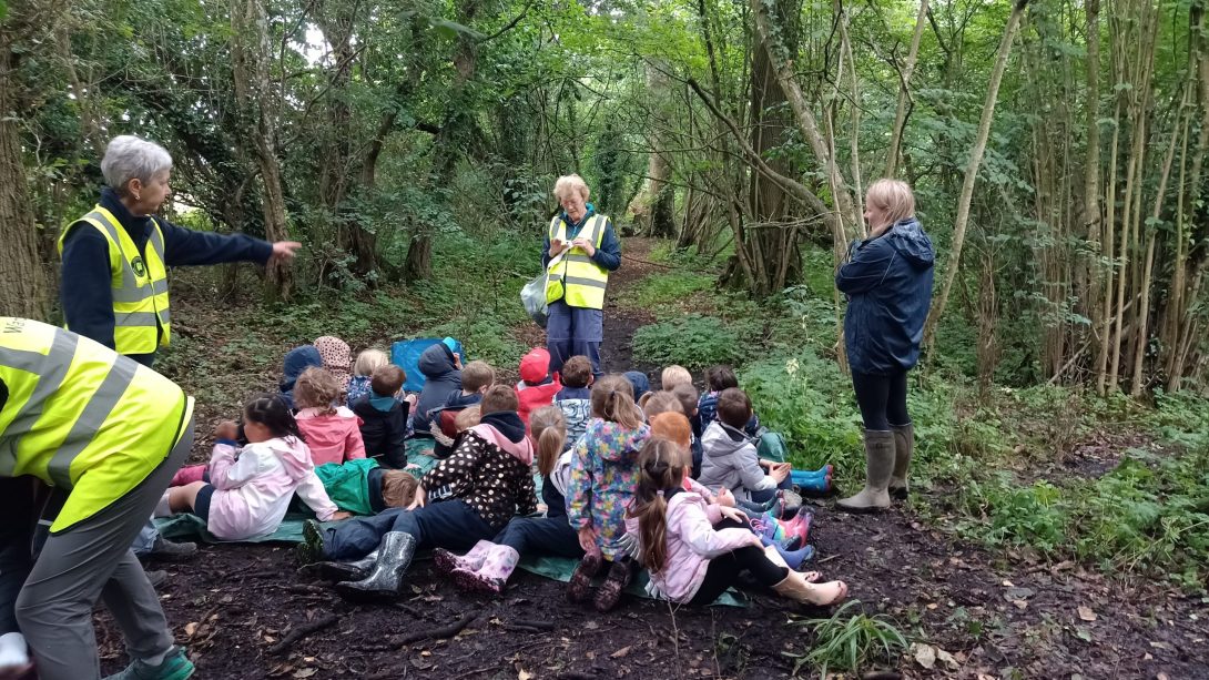 Photo of the children sat among trees in a wooded section of Greystones Nature Reserve, intently listening to one of the instructors