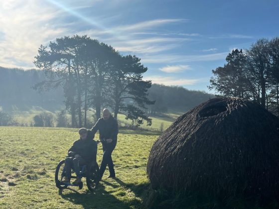 Images shows a man pushing a person in a Mountain Trike wheelchair across bumpy grass.