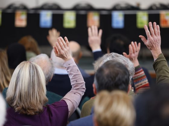 Image shows a conference room full of people - some have their hands up to ask a question.