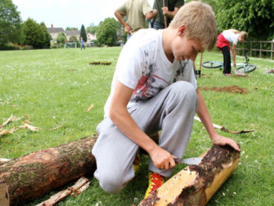 Image shows a young boy kneeling on grass chiselling a log.